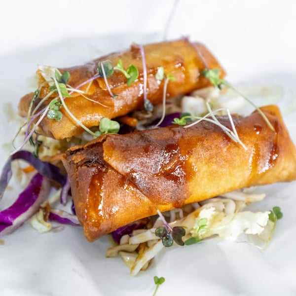 Two crispy vegan spring rolls with a sweet glaze, garnished with microgreens and shredded cabbage.