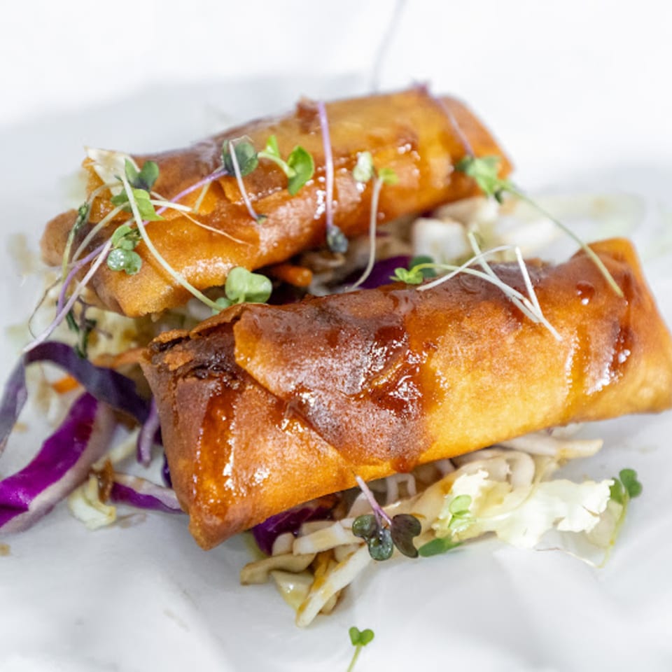 Two crispy vegan spring rolls with a sweet glaze, garnished with microgreens and shredded cabbage.
